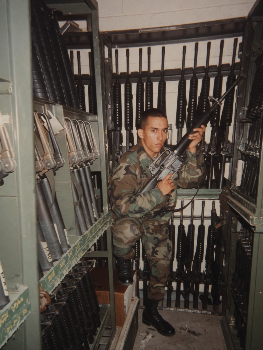 Commander as a young soldier in woodland camouflage kneeling in a Fort Leonard Wood armory weapons vault holding a service rifle